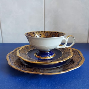 May include: A porcelain tea cup and saucer set featuring a deep cobalt blue and gold filigree pattern. The cup has a white interior with a gold rim and a pedestal base, while the matching saucer and plate display intricate gold scrollwork.