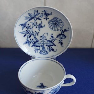 Op de afbeelding: Een witte porseleinen theekop en bijpassende schotel met een blauw uienpatroon. De schotel toont een gedetailleerd bloemen- en plantenontwerp in blauw, terwijl de kop een eenvoudig handvat en een klein blauw bloemmotief op de bodem heeft.