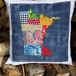 Puede incluir: Una almohada blanca con un inserto de tela vaquera azul que presenta un diseño de colcha de retazos del estado de Minnesota. La colcha está hecha de varias telas en rojo, blanco, azul, verde, amarillo y rosa.