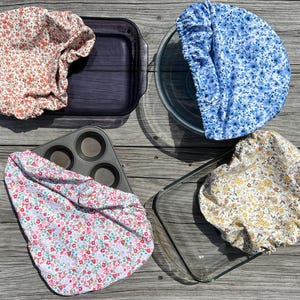 May include: Four floral fabric bowl covers in red, blue, pink, and yellow patterns. The covers are placed over a dark blue baking dish, a clear glass bowl, a muffin tin, and a clear glass baking dish. The covers are made of a soft, flexible fabric.