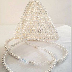 May include: A white triangular beaded bag with a long white beaded strap. The bag is made of small, round pearls and has a simple, elegant design.