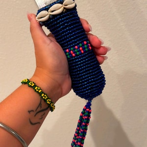 May include: A blue beaded handle adorns a machete blade. The handle is decorated with seashells, and a tassel of blue, green, and pink beads hangs from the bottom. The word "Thursday" is written in cursive.