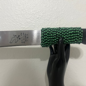 May include: A silver machete with a black and green beaded handle. The blade is engraved with the text "Gavilan De N. Colombia De Colp. K. F.K."