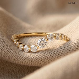 Könnte beinhalten: Ein geschwungener Goldring mit mehreren klaren Edelsteinen. Der Ring hat eine Reihe kleinerer Steine, die allmählich größer werden und in einem größeren zentralen Stein gipfeln. Vor einem neutralen Stoffhintergrund.