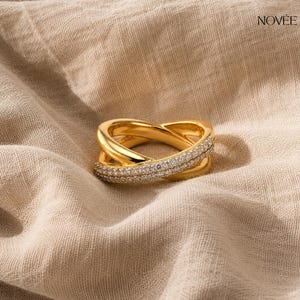 May include: A gold ring featuring a crossover design, embellished with small, glittering stones. The ring is displayed on a neutral-coloured fabric, with the brand name "NOVEE" visible in the upper right corner.
