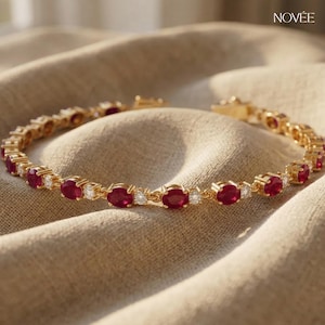 May include: A gold bracelet with oval-cut red gemstones and small, clear accent stones. The bracelet is displayed on a neutral fabric. The brand name "NOVÉE" is visible in the upper right corner.