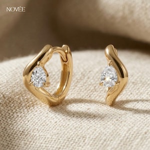 May include: A pair of gold hoop earrings, each featuring a pear-cut, clear gemstone. The earrings have an unusual, flowing design and are presented on a neutral fabric. The brand name "NOVEE" is visible in the upper left corner.