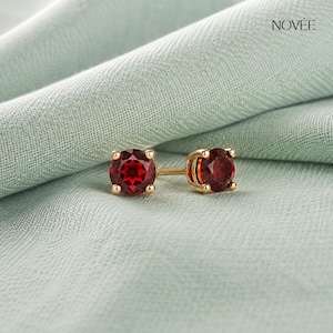 May include: A pair of round-cut red garnet stud earrings set in yellow gold prongs. The earrings feature a faceted gemstone design with a brilliant deep red hue. The word NOVÉE is printed in the upper right corner of the image.