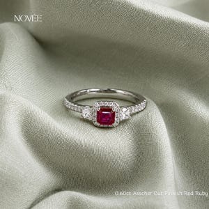 May include: A silver ring with a square, pinkish-red ruby center stone, surrounded by a halo of smaller diamonds. The band is also adorned with small diamonds. The ring is set against a soft, green fabric. Text on the image reads "0.60ct Asscher Cut Pinkish Red Ruby."