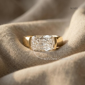 May include: A gold ring featuring a large, clear, cushion-cut diamond. The ring is displayed on a neutral fabric. The text reads "3.5 Carat Cushion Cut". The brand name "NOVEE" is visible in the upper right corner.