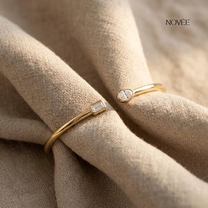 Könnte beinhalten: Ein goldfarbenes, offenes Armband mit zwei klaren, rechteckigen und oval geformten Edelsteinen. Das Armband ist vor einem neutralen Stoffhintergrund platziert. Das Wort "NOVEE" ist in Schwarz oben rechts aufgedruckt.