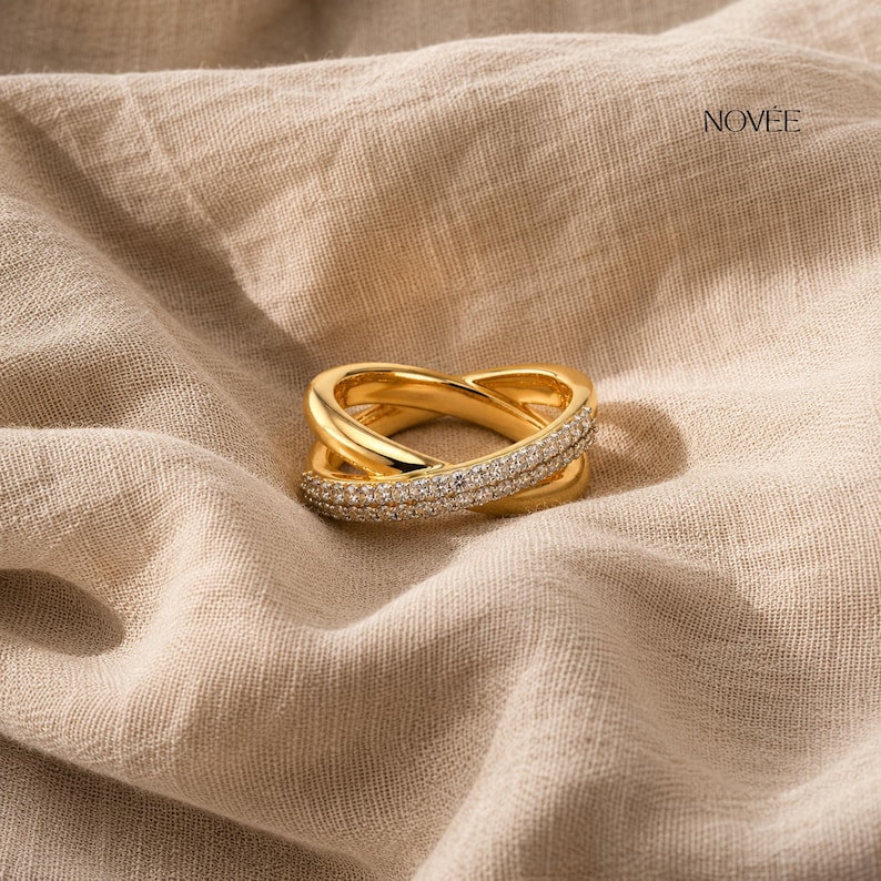 May include: A gold ring featuring a crossover design, embellished with small, glittering stones. The ring is displayed on a neutral-coloured fabric, with the brand name "NOVEE" visible in the upper right corner.
