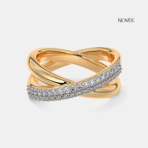May include: A gold ring with a crossover design. The ring has a band of small, sparkling white stones. The ring has a polished finish and is set against a neutral background. The brand name "NOV&Eacute;E" is visible. The ring is a piece of jewellery.