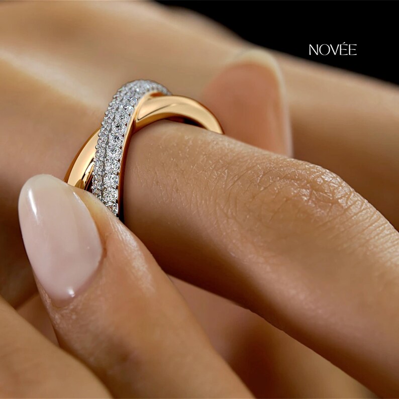 May include: A gold and diamond ring with a crossover design. The ring features a band of gold that crosses over a band of small, sparkling diamonds. The ring is on a person's finger. The brand name "NOV&Eacute;E" is visible in the background.