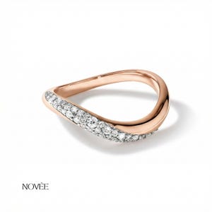 Puede incluir: Anillo de oro rosa con diseño ondulado, con una fila de diamantes blancos brillantes en un lado. El anillo tiene un acabado liso y pulido y se presenta sobre un fondo blanco. La marca "NOVEE" está impresa en la parte inferior.
