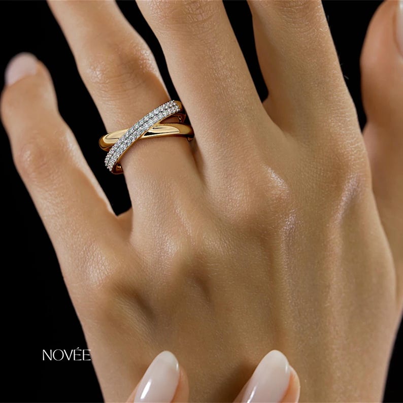 May include: A gold ring with a crisscross design, adorned with small, sparkling clear stones. The ring is on a person's finger, with the brand name "NOV&Eacute;E" visible. The background is black.