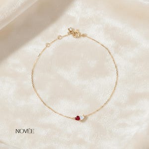 May include: Delicate gold bracelet featuring a small red heart-shaped gemstone and a white heart-shaped gemstone. The bracelet has a delicate chain and a small clasp. The brand name "NOVÉE" is printed in black on the lower left.