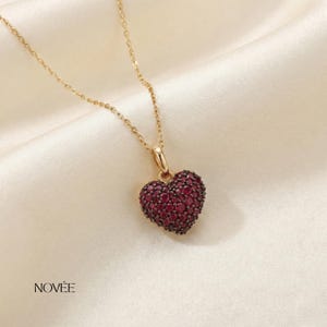 Puede incluir: Un collar dorado con un colgante en forma de corazón. El colgante está cubierto de pequeñas piedras preciosas de color rojo oscuro. El collar se muestra sobre una tela blanca satinada. La marca "NOVEE" está impresa en negro.
