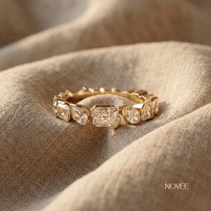 Puede incluir: Un anillo de oro con múltiples piedras preciosas claras y brillantes de varias formas, incluyendo cortes de pera y esmeralda. El anillo se encuentra sobre un fondo de tela de tonos neutros, con la marca "NOVÉE" visible en la esquina inferior derecha.