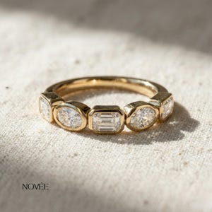 May include: A gold ring with a series of clear gemstones. The ring has a polished finish and is set with a mix of oval and emerald-cut stones. The ring is displayed on a neutral-coloured fabric.