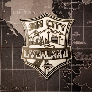Puede incluir: Un parche blanco y negro con el texto "Sin City Overland" y un gráfico de un horizonte de ciudad con montañas y una carretera. El texto "Las Vegas" también está incluido en el gráfico.