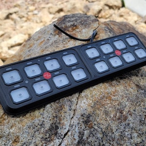 May include: A black, rectangular, multi-button controller with a red button labeled "OFF/ON". The controller has 12 buttons arranged in two rows of six. The controller is sitting on a rock.