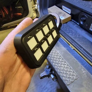 May include: A black rectangular panel with 10 white buttons. The panel is held in a person's hand.