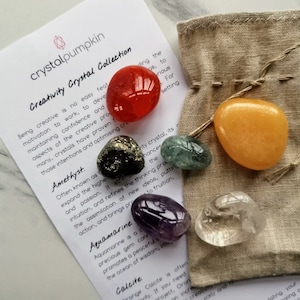 May include: A set of seven crystals in a small cloth bag. The crystals are amethyst, aquamarine, calcite, clear quartz, citrine, pyrite, and red aventurine. The crystals are arranged in a circle around a small card with the text "Creativity Crystal Collection".