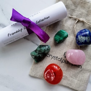 Könnte beinhalten: Ein kleiner Beutel mit einem violetten Band darum gebunden, der ein Set aus sechs getrommelten Kristallen enthält. Die Kristalle sind grün, blau, rosa und rot. Der Beutel ist mit "crystalpumpkin" beschriftet. Das Band ist um ein aufgerolltes Stück Papier gebunden, auf dem der Text "Friendship Collection of our everyday" steht.
