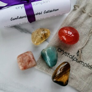 May include: A set of six tumbled crystals in a small cloth bag. The crystals are yellow, orange, red, green, brown, and peach. The bag has the text "crystalpumpkin" printed on it. The crystals are part of a "Confidence Crystal Collection".
