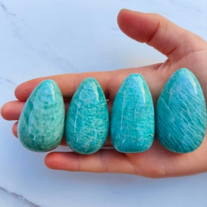 Peut inclure: Quatre œufs en cristal d'amazonite lisses et verts, chacun avec un motif unique de lignes tourbillonnantes, sont présentés dans une main.