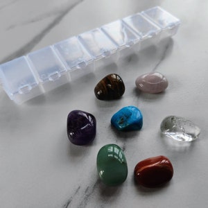 Op de afbeelding: Een transparante plastic opbergdoos met zeven vakken, gevuld met zeven verschillende gekleurde edelstenen. De edelstenen omvatten groen, blauw, paars, bruin, roze, helder en rood.