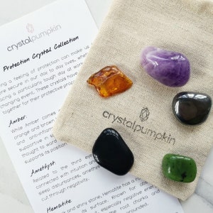 May include: A set of five crystals in a small, beige cloth bag. The crystals are amber, amethyst, hematite, black obsidian, and green jade. The bag has the text "crystalpumpkin" printed on it.