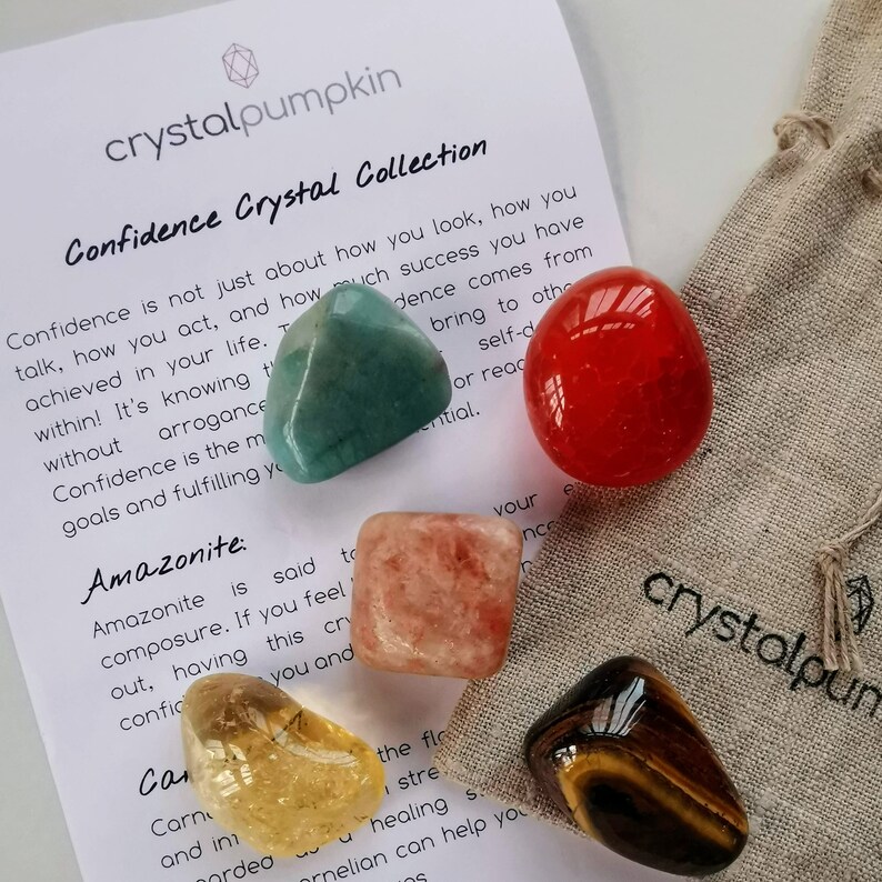 May include: A set of six tumbled crystals for confidence, including amazonite, carnelian, clear quartz, red jasper, rose quartz, and tiger's eye. The crystals are arranged on a white background with text about confidence.
