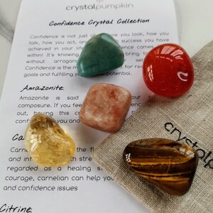 May include: A set of five tumbled crystals in a small pouch. The crystals are green, orange, red, yellow, and brown. The pouch is made of natural fabric and has the word "crystal" printed on it. The crystals are labeled with their names: Amazonite, Carnelian, Citrine, Tiger's Eye, and Aventurine.