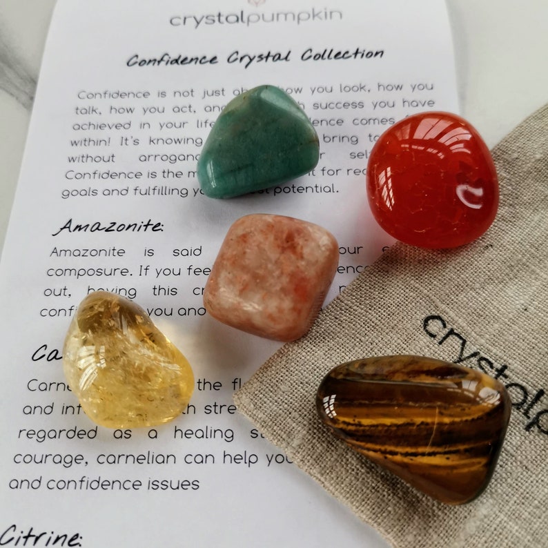 May include: A set of five tumbled crystals in a small pouch. The crystals are green, orange, red, yellow, and brown. The pouch is made of natural fabric and has the word "crystal" printed on it. The crystals are labeled with their names: Amazonite, Carnelian, Citrine, Tiger's Eye, and Aventurine.