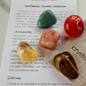 May include: A set of six tumbled crystals for confidence, including amazonite, carnelian, citrine, clear quartz, sunstone, and tiger's eye. The crystals are arranged on a beige fabric pouch with the text "Confidence Crystal Collection" and descriptions of each crystal.
