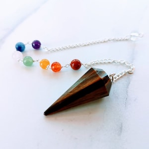 Op de afbeelding: Een donkerbruin houten slinger met een zilveren ketting en een regenboog van chakra-parels. De parels zijn in de volgorde van de chakra's: wortel, heiligbeen, zonnevlecht, hart, keel, derde oog en kroon.