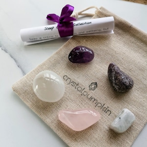 Könnte beinhalten: Eine kleine, beige Stofftasche mit einem violetten Band und einer aufgerollten Notiz, auf der steht: "Schlaf-Kollektion, Schlaf ist entscheidend für Gesundheit und Wohlbefinden, aber..." Die Tasche enthält fünf getrommelte Kristalle: Amethyst, klarer Quarz, Rosenquarz, Amethyst und Mondstein.