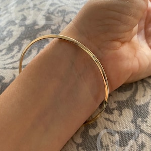 Puede incluir: Un brazalete de color dorado. El brazalete es una banda fina y sencilla que se curva alrededor de la muñeca. El metal tiene un acabado brillante. El fondo es una tela estampada.