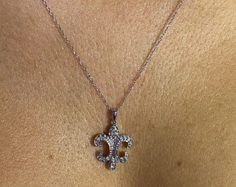 May include: A silver necklace featuring a fleur-de-lis pendant encrusted with small, sparkling stones. The pendant hangs from a delicate chain, creating a classic and elegant look. The necklace is shown against a skin tone background.