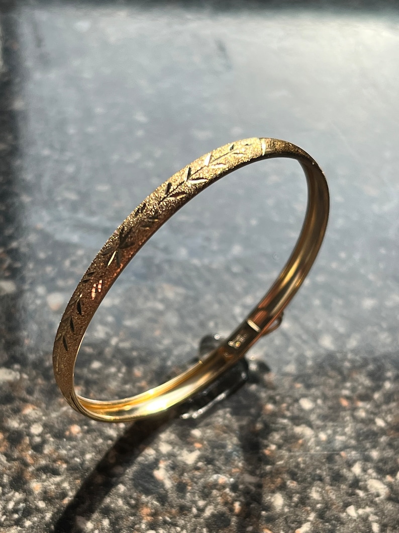 Puede incluir: Un brazalete de oro con un dise&ntilde;o texturizado. El brazalete est&aacute; hecho de un metal liso y brillante y tiene un dise&ntilde;o simple y elegante.