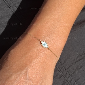 Puede incluir: Una delicada pulsera dorada con un dije en forma de ojo con iris azul claro y fondo blanco. La pulsera se lleva en la muñeca, mostrando la fina cadena y el diseño del dije. La joya es elegante y minimalista.