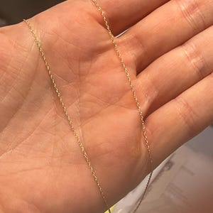 May include: A delicate gold-colored chain necklace with a twisted rope design. The necklace is shown against a neutral background, highlighting its fine details and subtle shine. The chain is lightweight and appears to be a classic jewelry piece.