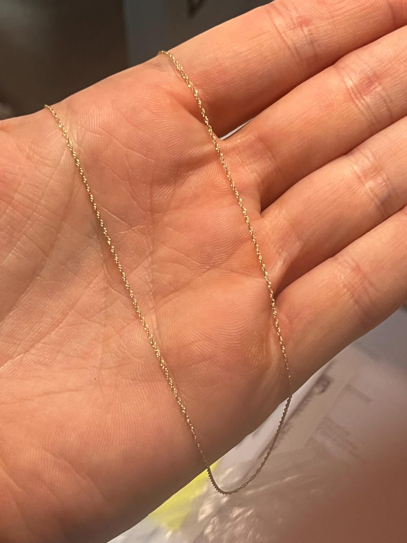May include: A delicate gold-colored chain necklace with a twisted rope design. The necklace is shown against a neutral background, highlighting its fine details and subtle shine. The chain is lightweight and appears to be a classic jewelry piece.