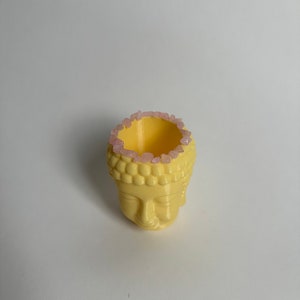 May include: A yellow ceramic planter shaped like a Buddha head with pink crystals around the rim.