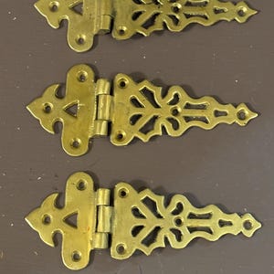 May include: Three ornate brass hinges with decorative cut-out designs. Each hinge has a triangular shape on one end and a detailed, elongated shape on the other, connected by a central hinge. The brass has a warm, golden hue.