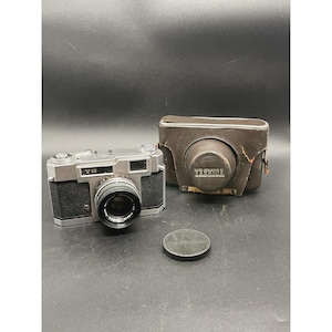 May include: A silver Yashica camera with a black lens and a brown leather case. The camera is sitting on a gray surface with a black lens cap next to it.