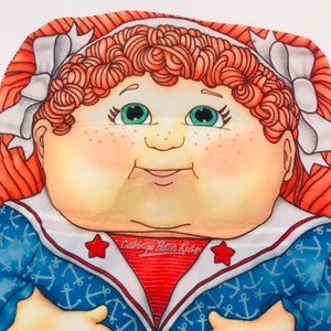 May include: A Cabbage Patch Kids doll with red hair, green eyes, and a sailor outfit. The doll is wearing a blue and white sailor shirt with anchors and a red and white striped collar. The doll's face is painted with rosy cheeks and a sweet smile. The doll's name is written on the shirt: "Cabbage Patch Kids".