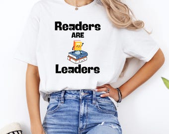 May include: White t-shirt with the text "Readers are Leaders" in black. The shirt features a graphic of stacked books with smiling faces and a book on top. The model is wearing blue jeans.